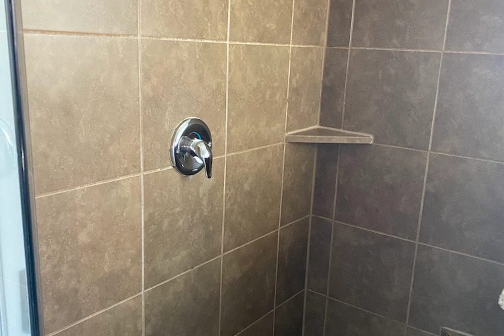A walk-in shower with brown ceramic wall tile, a chrome valve and a corner shelf